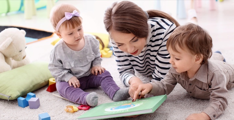 Tagespflegeperson schaut mit zwei Kleinkindern ein Bilderbuch an